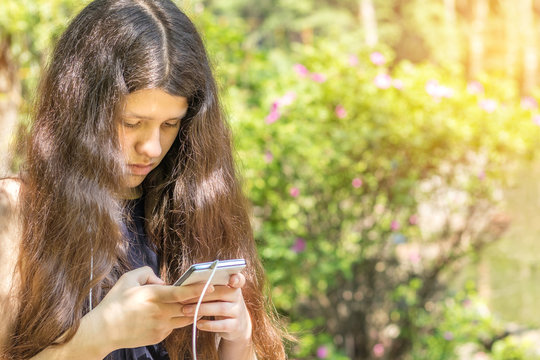 Young long haired girl using the phone in the city park on a summer sunny day. Walking in summer park on the summer day. Portrait female  teenager with copy space.