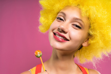 laughing girl in a bright orange wig on a pink background holds in hands chupa chups