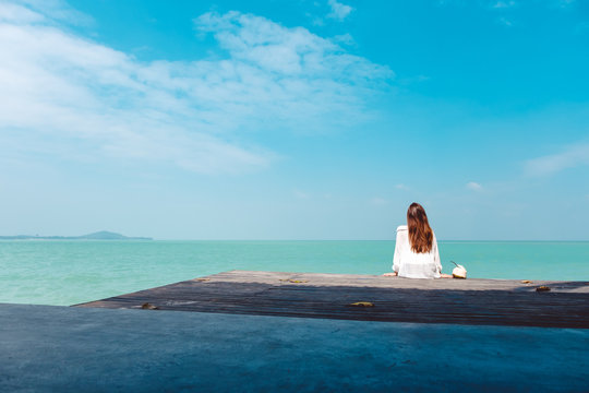 A Beautiful Asian Woman On White Dress Sitting And Looking At The Sea And Blue Sky On Wooden Balcony With Feeling Relaxed