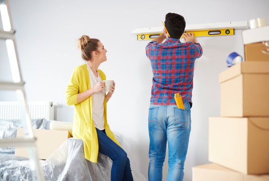 Young couple at home, surrounded by cardboard boxes, man checking shelf with spirit level