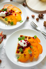 Plates of Belgian waffles with persimmon, pomegranate seeds and sour cream on white table cloth