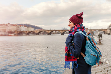 Young woman tourist in coat with backpack standing near bridge over the Vltava river in Prague