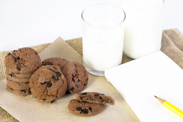Chocolate chip cookies on brown paper and sack cloth with glass of milk and notepad on white background. With copy space for text