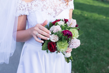 Beautiful wedding colorful bouquet in hands of the bride. Trendy and modern wedding flowers. A wedding ring on the finger of the bride. Woman in wedding dress outdoors. Green background.
