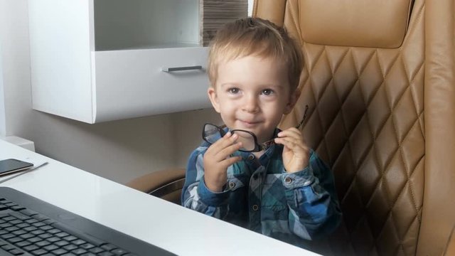 4k footage of adorable smiling toddler boy sitting in big boss chair at office and wearing eyeglasses. Concept of little businessman