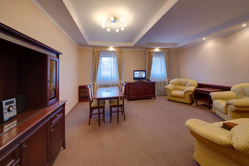 A light drawing room with upholstered furniture, a dining table, chairs, a sideboard and the TV set on a bedside table