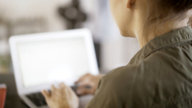 A young woman wearing a green shirt typing at her laptop in an office. Over the shoulder shot. Blurred laptop screen.
