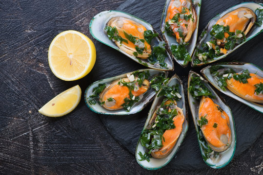 Above view of boiled green mussels with parsley and lemon on a stone slate tray, dark brown stone background, horizontal shot