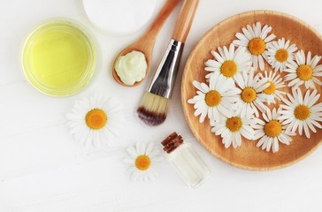 Holistic homemade herbal cosmetics with camomile, essential oils, bowls, facial cream, top view white wooden table.