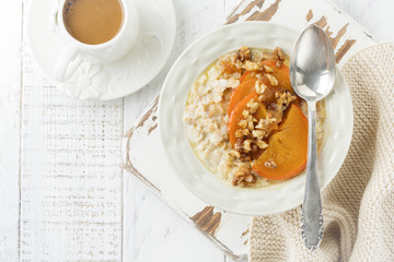 Oatmeal porridge with caramelized persimmon and walnuts in a light ceramic bowl on a white wooden background. Selective focus. Top view. Copy space.