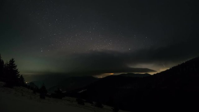 Starry night sky with stars and low clouds moving over mountains. Time lapse zoom in