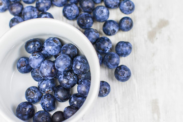 Juicy blueberries in a small white jar with scattered blueberries on the wooden table.