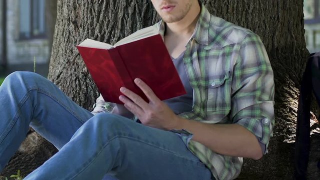 Guy reading gripping book under tree, engrossed into plot, avid book reader