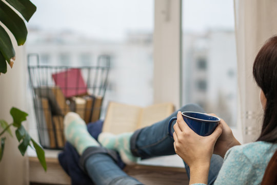 Young Woman Sitting In A Chair Looking Out The Window, Relaxing, Reading A Book And Drinking Coffee Or Tea.