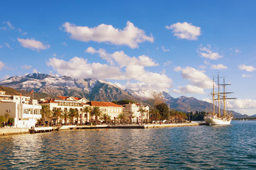 Fototapeta na wymiar Snowy mountains and green seaside town under blue sky with white clouds. Montenegro, winter. View of embankment of Tivat city