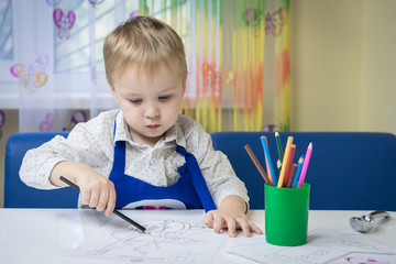 cute pensive caucasian boy 3 years old drawing with colored pencil in a notebook sitting at a desk in a classroom in a kindergarten (play room)