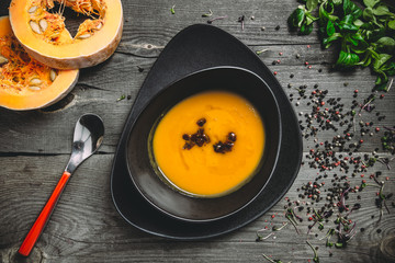 Pumpkin cream soup with dried tomatoes, basil and spices, in a round black plate on a dark wooden background, beside is a spoon