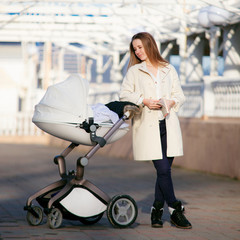 A young mother in a white coat walks along the seafront with a child in a stylish stroller. Concept of active motherhood.