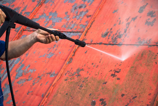 Man Is Washing The Roof With A High Pressure Washer. Worker Cleaning A Red Metal Sheet Roof By Water. Detail 