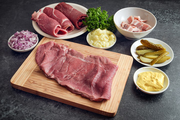 Preparing beef roulades with the ingredients of mustard, onions, pickled cucumbers, bacon and fresh meat on a wooden cutting board and a dark kitchen countertop