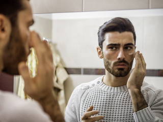 Handsome young man applying moisturizing cream on face in front of home bathroom mirror