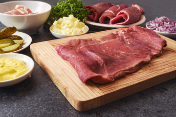 cooking beef roulades with the ingredients as mustard, onions, pickled cucumbers, bacon and the fresh meat on a wooden cutting board and a dark kitchen countertop