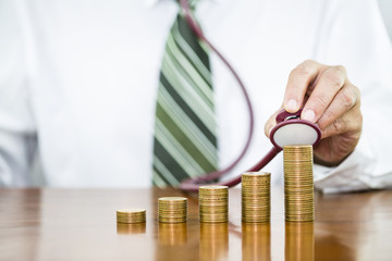 Businessman hand holding stethoscope checking stack of money coins arranged as a graph on wooden table, concept of financial health check and saving money