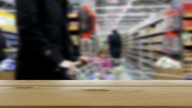 Top of a wooden table in the foreground. Defocused image of a supermarket and buyers in the background.