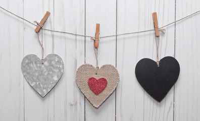 Three hearts, a silver one, a black one and a brown one with a smaller red heart in it's middle hanging on a line in front of a white washed wooden fence