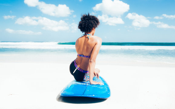 Surfer girl with afro hairstyle sitting on blue longboard on the white sand in front of a breathtaking seascape, looking at waves at Dreamland beach, Bali, Indonesia	