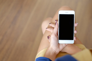 Mockup image of woman's hand holding white mobile phone with black screen on thigh with wooden floor background in modern
