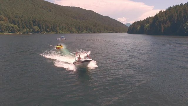 Aerial tracking shot of speedboat pulling women and kid on a colorful inner tube across lush treelined lake