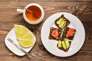 Snack with red fish and avocado on rye crackers. Tea with lemon. Wooden rustic background.
