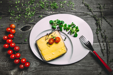 Delicious sandwich with bacon and cheese under the béchamel sauce with cherry tomatoes and spinach. White round dish, branch of tomatoes cherry, thyme and fork on gray wooden background