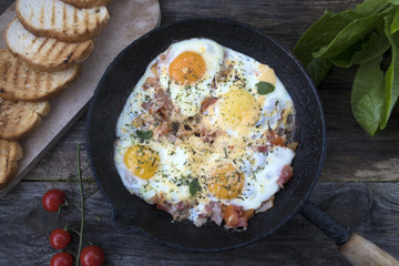 Fried eggs in an old frying pan, tomatoes, lettuce leaves and fried bread on a wooden background.
