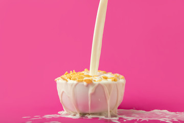 pouring fresh healthy milk into bowl with corn flakes on pink