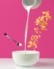 Fototapeta na wymiar pouring fresh healthy milk from jug to bowl, spoon and corn flakes on pink