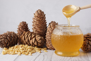 Honey is poured from a wooden spoon into a glass keg, near cedar cones and nuts on a light table