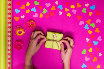 Valentines day gift wrapping with boxes over pink background and colorful paper hearts around. Process of handmade Present. Creative diy craft hobby. Holiday decorations