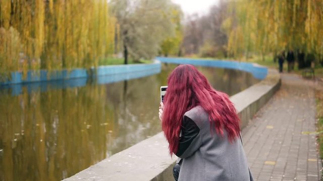 Young attractive woman with red hait taking picture on her smartphone while standing by an artificial pond in an autumn park