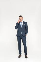 full length view of stylish young businessman standing with hand in pocket and talking on smartphone isolated on grey