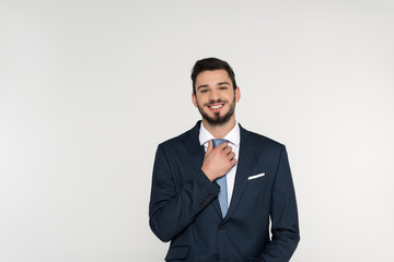 handsome young businessman adjusting necktie and smiling at camera isolated on grey