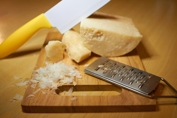 3 different pieces of coarse Italian Parmesan cheese, a special cheese knife for Parmesan, enters the cheese head at an angle, a small iron grater for cheese, grated parmesan flakes on a wooden board.