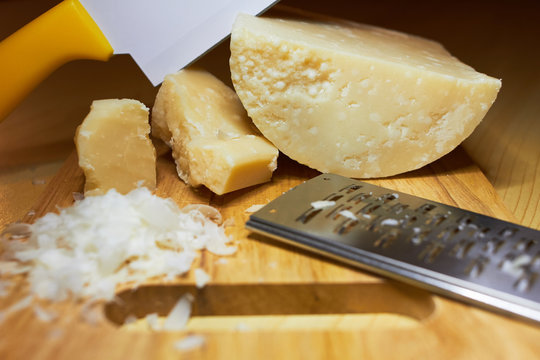 3 different pieces of coarse Italian Parmesan cheese, a special cheese knife for Parmesan, enters the cheese head at an angle, a small iron grater for cheese, grated parmesan flakes on a wooden board.