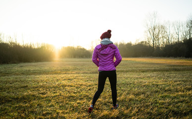 A young active lady walking through a frosty winter field towards a group of trees with the golden light of sunset brightening up the sky