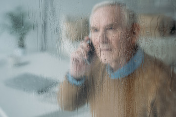 Behind the rainy glass view of senior disturbed man making a phone call