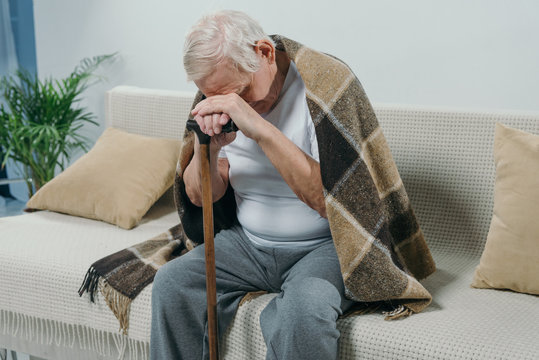 Tired senior man wearing plaid leans on a cane while sitting on sofa