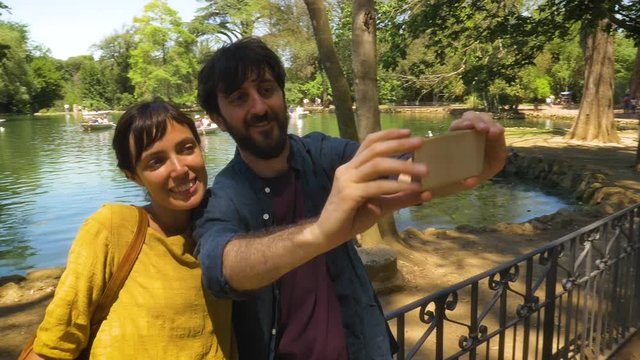 Young happy Couple Taking Selfies with smartphone by a Pond in Villa Borghese Park in Rome on sunny summer day yellow dress smiling slow motion steadycam