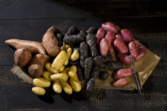 Different sorts of potatoes and peeler on brown paper, jute and dark wood