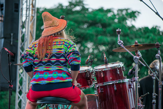 Shot of a girl with colorful clothes and orange hat playing drums on stage during music festival in summer. Band performing their songs.